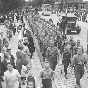 Black and white photo of Nazi Storm Troopers marching through the city streets of Berlin in 1932 as civilians walk along the sidewalk or wait to cross the street until the Nazis have passed.