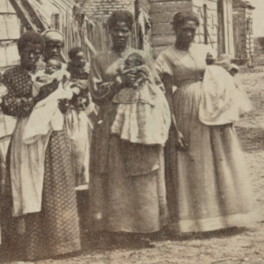 A black and white photo of four enslaved women standing together outside their cabins holding their infants. South Carolina, 1860s.