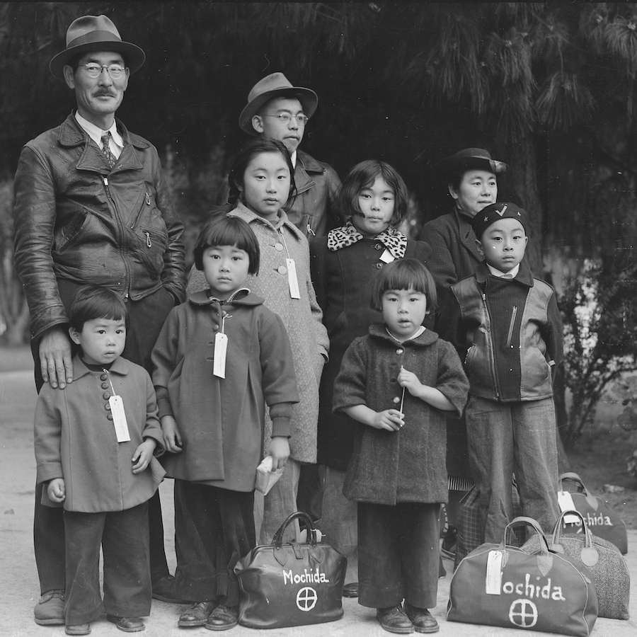 Black and white photo of a Japanese American family of eight standing together with their bags to be taken to a U.S. concentration camp during WWII. The children have large tags on them.