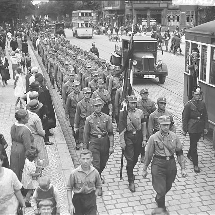 Black and white photo of Nazi Storm Troopers marching through the city streets of Berlin in 1932 as civilians walk along the sidewalk or wait to cross the street until the Nazis have passed.